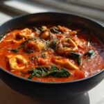 A close-up of a dark bowl filled with rich, creamy tomato tortellini soup, featuring spinach and herbs.