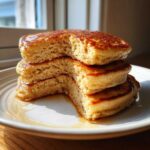 Close-up of a stack of fluffy Gluten-Free Buttermilk Pancakes with a piece removed, drizzled with syrup.