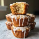 A stack of Glazed Lemon Poppy Seed Muffins, with the top muffin having a bite taken out to show the moist interior.