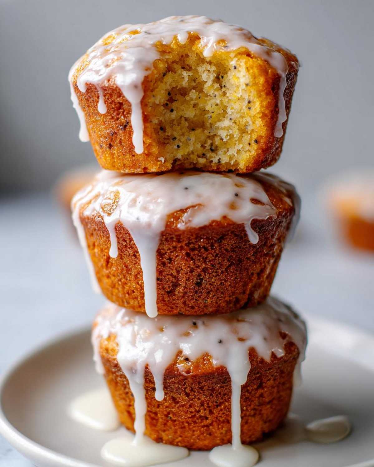 Close-up of three Glazed Lemon Poppy Seed Muffins stacked, with the top one having a bite taken out showing the poppy seeds inside.
