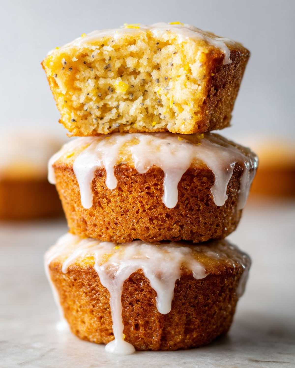 Close-up of three Glazed Lemon Poppy Seed Muffins stacked, with the top one broken open showing poppy seeds.