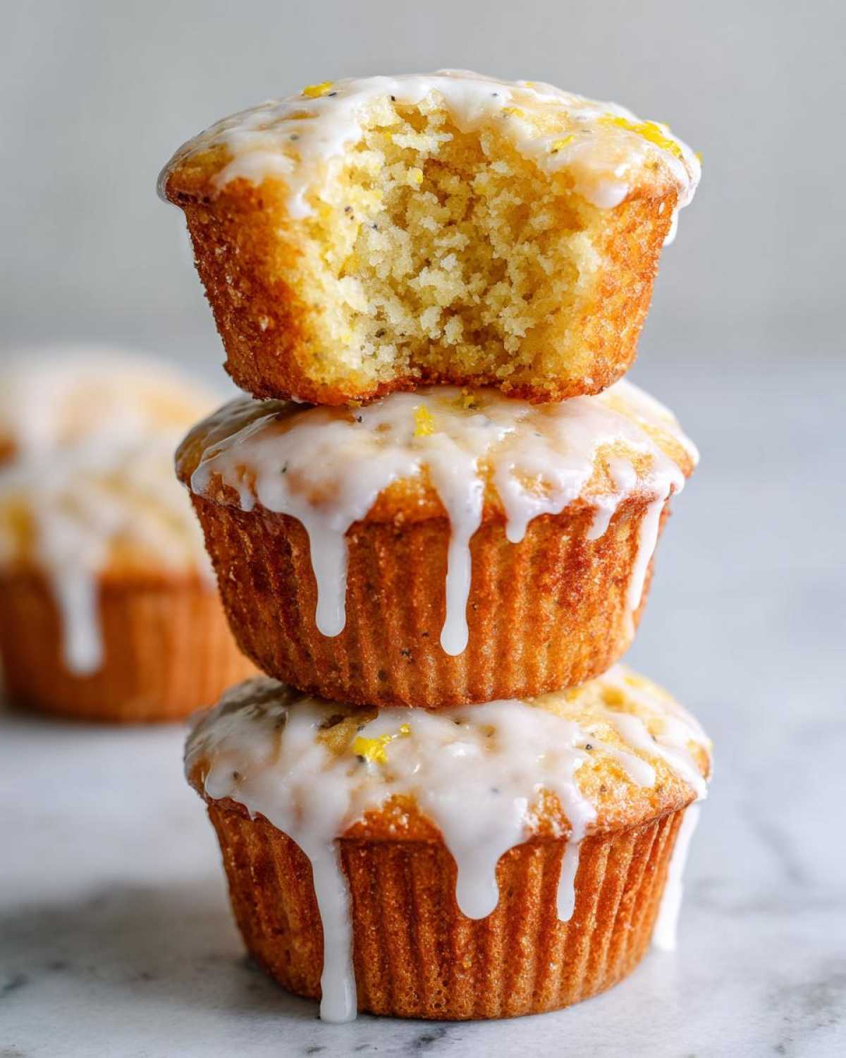 A stack of three Glazed Lemon Poppy Seed Muffins, with the top one having a bite taken out to show the texture.