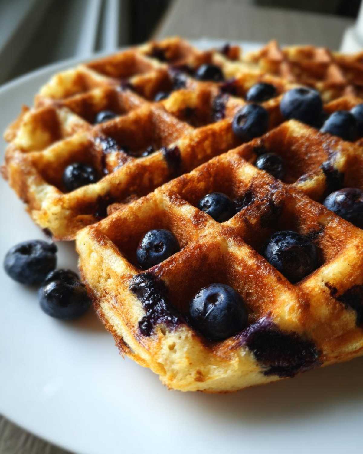 Close-up of golden brown Gluten-Free Blueberry Waffles topped with fresh blueberries on a white plate.