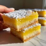 Two stacked Gluten-Free Lemon Bars topped with bright yellow curd and a dusting of powdered sugar, held by a hand.