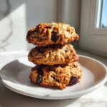 Three golden brown Gluten-Free Oatmeal Raisin Cookies stacked on a white plate near a window.