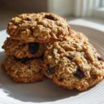 A stack of four golden-brown Gluten-Free Oatmeal Raisin Cookies piled on a white, ridged plate.