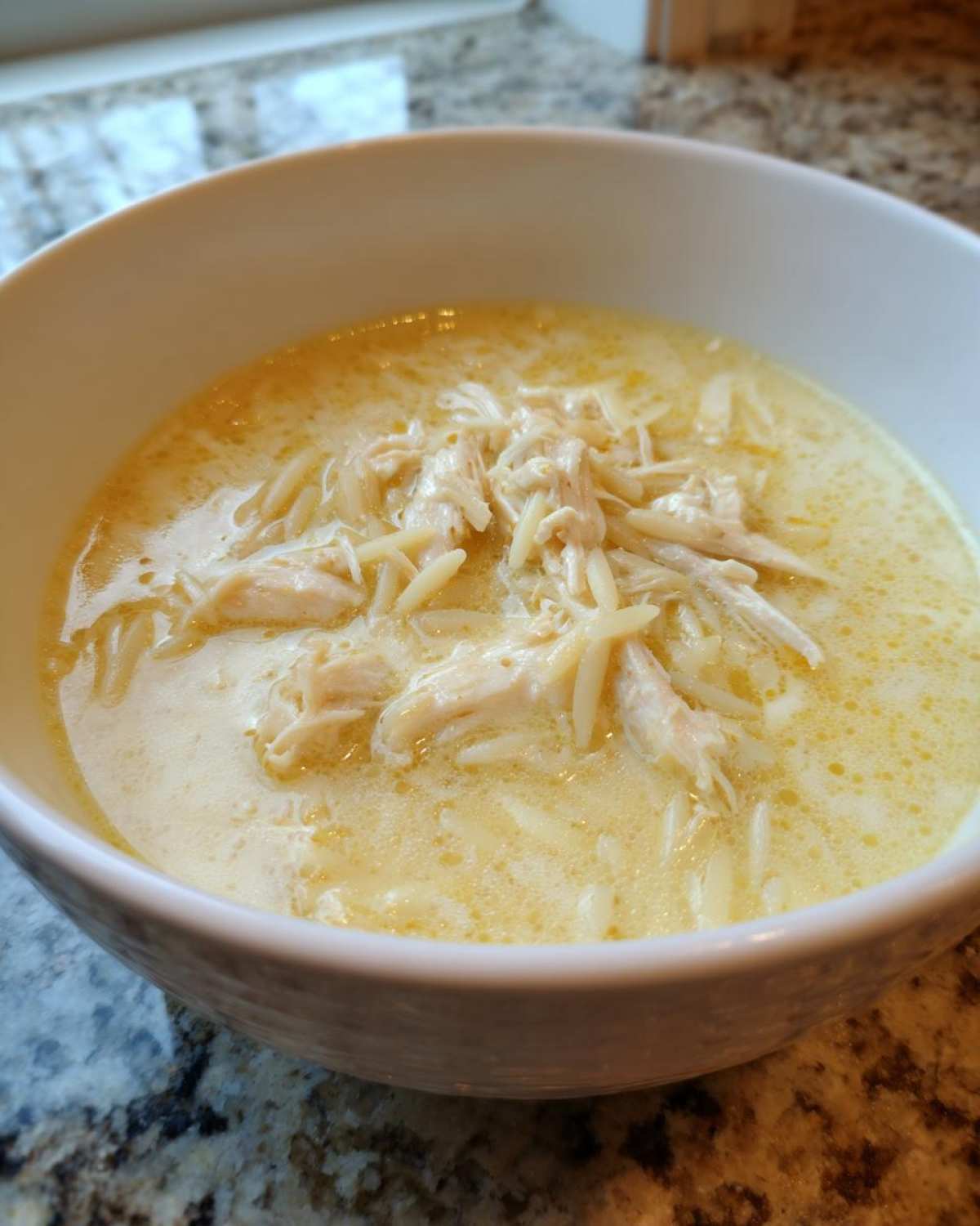 Close-up of a white bowl filled with creamy, yellow Greek Lemon Chicken Soup featuring shredded chicken and orzo pasta.