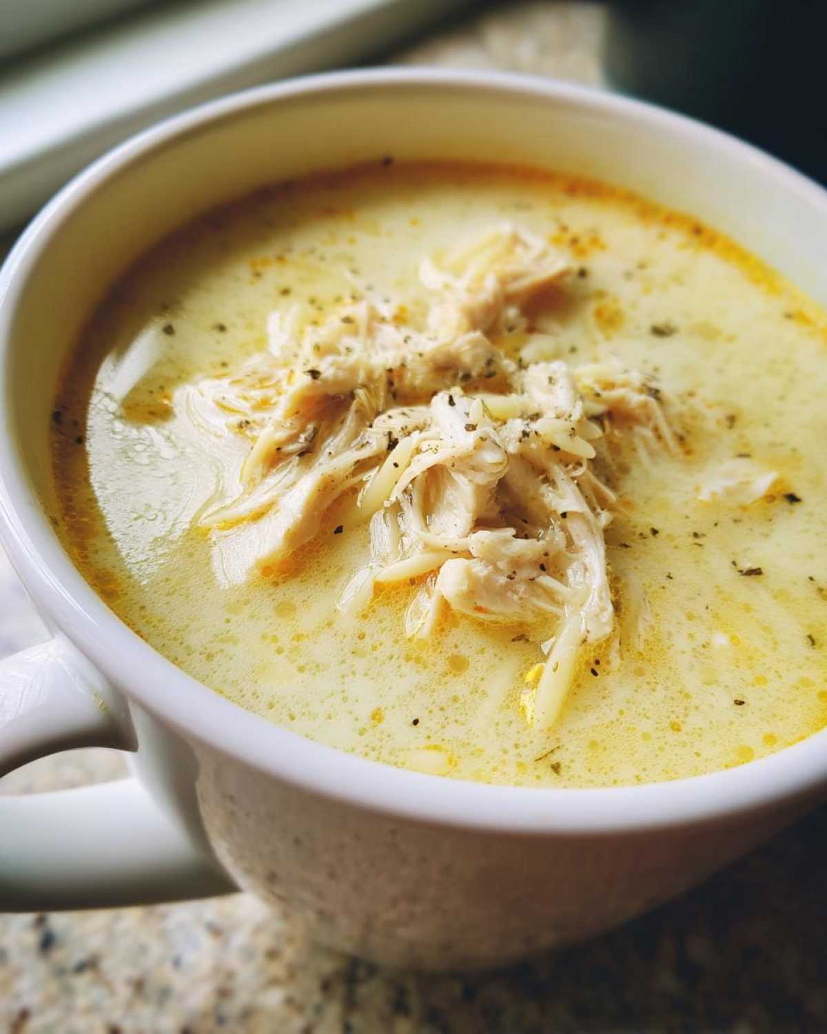 Close-up of a creamy, yellow bowl of Greek Lemon Chicken Soup topped with shredded chicken and black pepper.