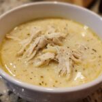 Close-up of a white bowl filled with creamy Greek Lemon Chicken Soup, topped with shredded chicken and herbs.