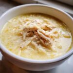 Close-up of a white bowl filled with creamy Greek Lemon Chicken Soup, topped generously with shredded chicken and orzo pasta.