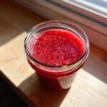 Close-up of bright red Homemade Strawberry Lemonade Concentrate with foam on top, sitting in a glass jar in sunlight.