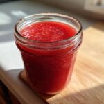 A glass jar filled with bright red Homemade Strawberry Lemonade Concentrate sitting on a wooden counter.