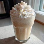 A tall glass of Homemade Vanilla Bean Frappuccino topped high with whipped cream and a sprinkle of spice.