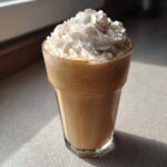 A tall glass of Homemade Vanilla Bean Frappuccino, topped generously with whipped cream and a sprinkle of spice.