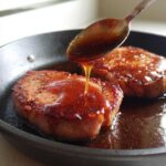 A spoonful of thick honey glaze is being poured over a seared Honey Glazed Ham Steak in a black skillet.