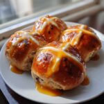 Four freshly baked Hot Cross Buns with Orange Glaze drizzled over the white icing crosses, sitting on a white plate.