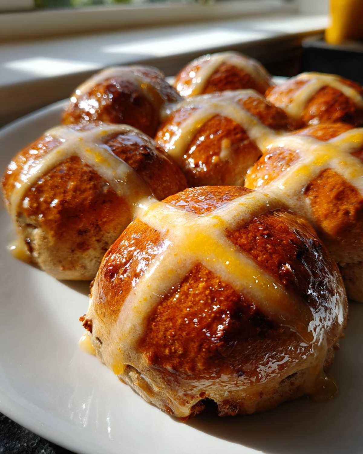 A close-up of several freshly baked Hot Cross Buns with Orange Glaze glistening on a white plate.