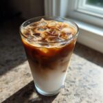 A tall glass of layered Iced Brown Sugar Oatmilk Latte with ice cubes, sitting on a countertop in sunlight.