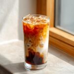 A tall glass featuring a beautiful swirl of dark coffee and white oatmilk in an Iced Brown Sugar Oatmilk Latte, topped with ice.