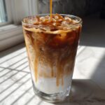 A tall glass being filled with espresso and caramel syrup over ice and milk to make an Iced Caramel Macchiato.