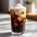 Close-up of a tall glass containing a rich Iced Mocha with ice, showing cream swirling into the dark coffee, topped with whipped cream.