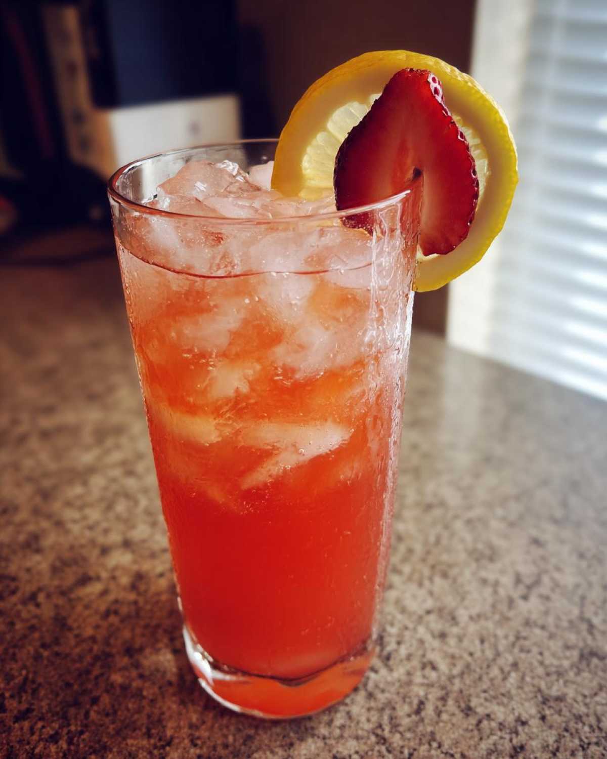 A tall glass filled with bright red Iced Strawberry Lemon Tea, ice cubes, and garnished with a strawberry slice and lemon wheel.