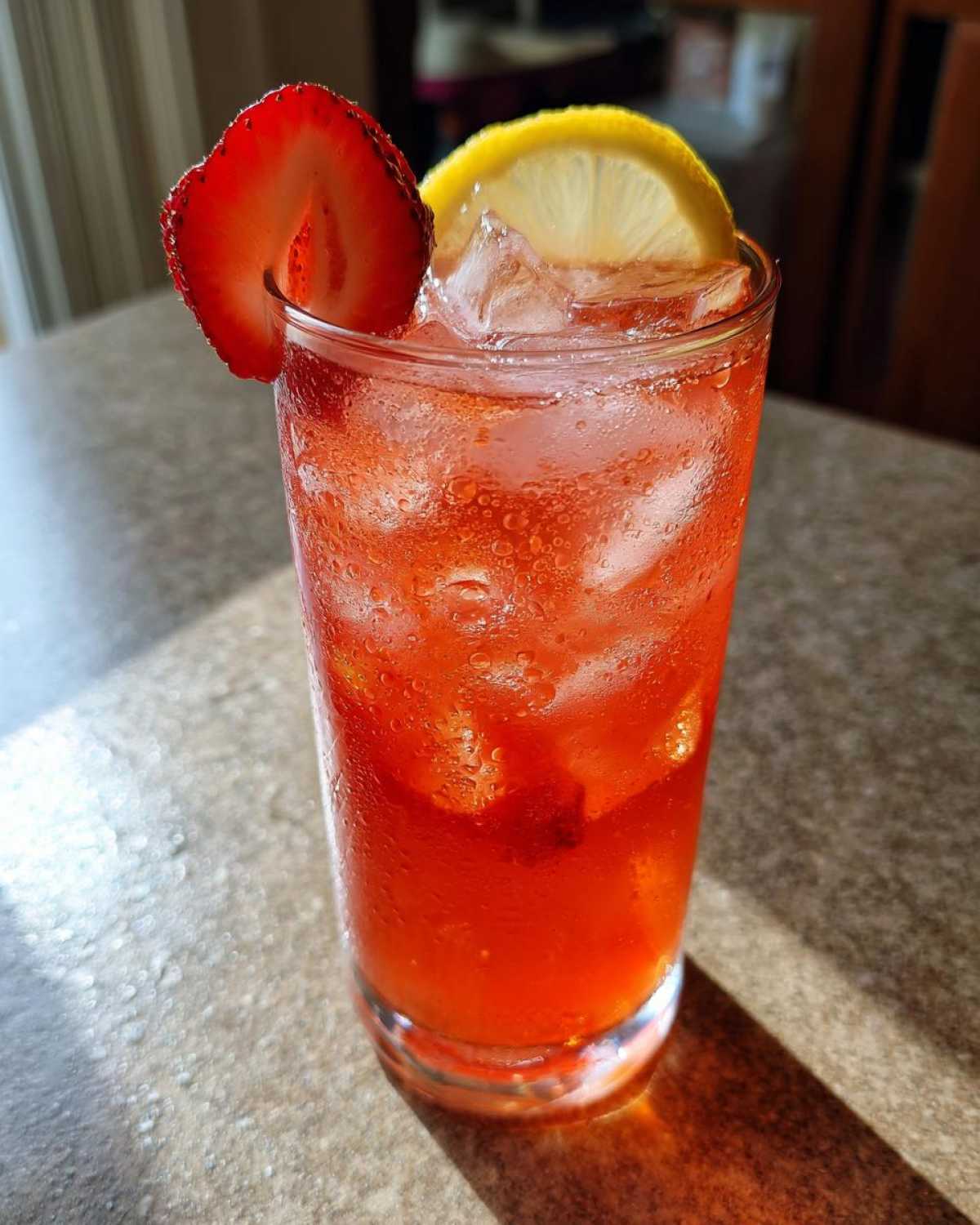 A tall, sweating glass filled with bright red Iced Strawberry Lemon Tea, ice, and garnished with a strawberry slice and lemon wheel.