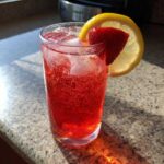 A tall glass filled with vibrant red Iced Strawberry Lemon Tea, ice cubes, and garnished with a fresh strawberry slice and lemon wheel.