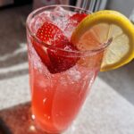 Close-up of a tall glass filled with bright pink Iced Strawberry Lemon Tea, ice, strawberries, and a lemon slice garnish.