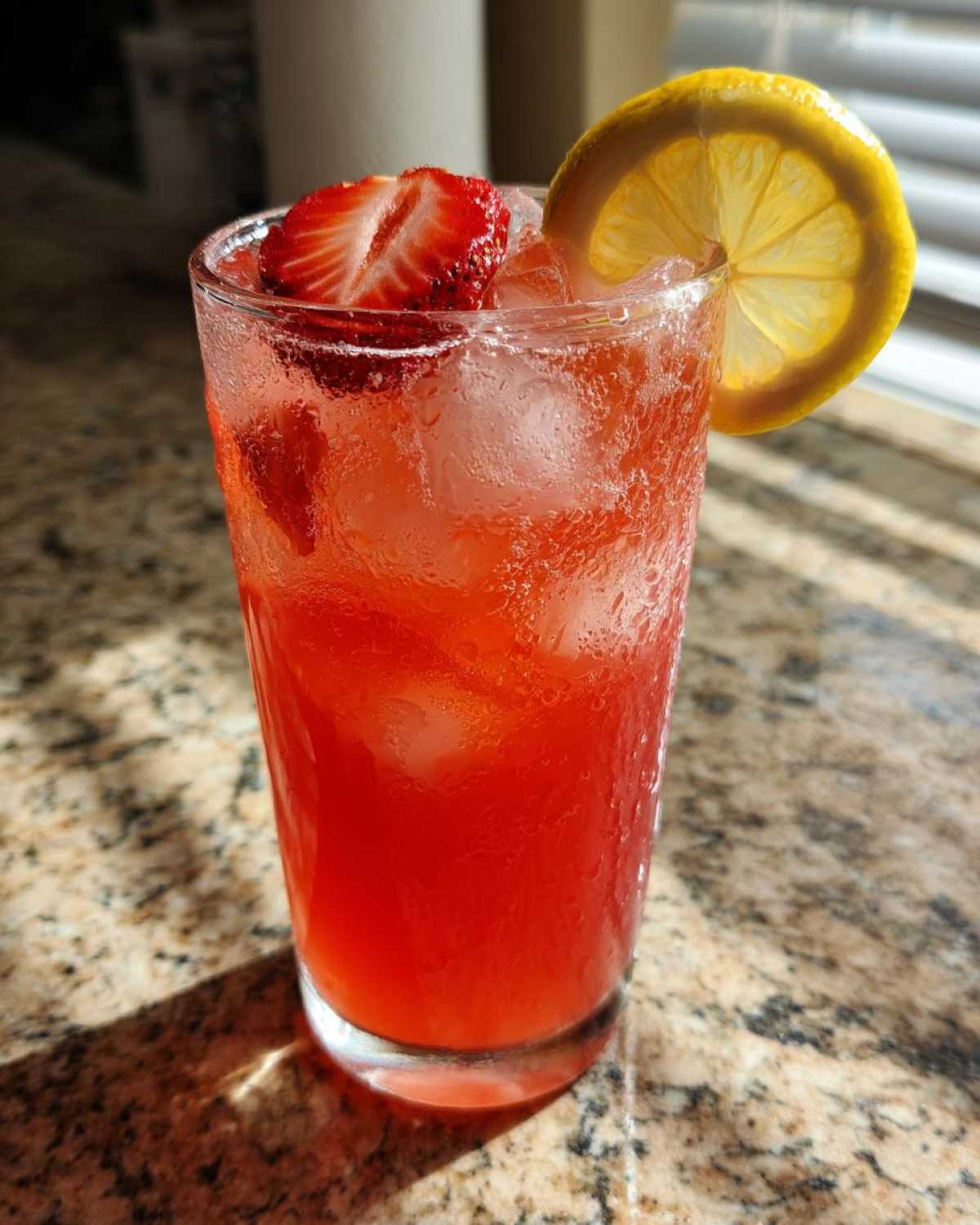 A tall, sweating glass filled with bright red Iced Strawberry Lemon Tea, ice, and garnished with a lemon slice and strawberries.