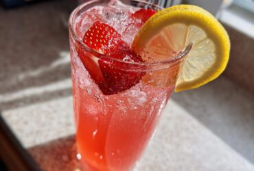 Close-up of a tall glass filled with bright pink Iced Strawberry Lemon Tea, ice, strawberries, and a lemon slice garnish.