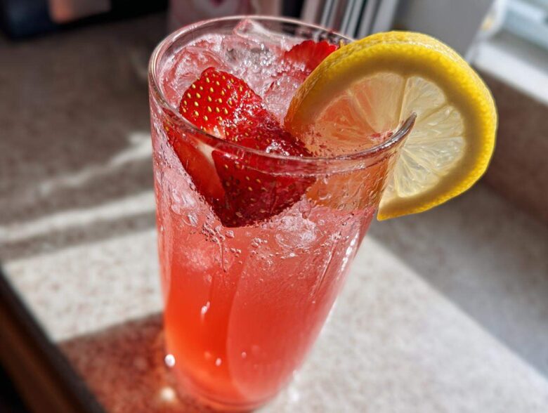 Close-up of a tall glass filled with bright pink Iced Strawberry Lemon Tea, ice, strawberries, and a lemon slice garnish.