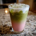 A tall glass showing a layered Iced Strawberry Matcha Latte with pink strawberry base, green matcha, and ice.