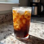 Close-up of a tall glass filled with Iced Vanilla Chai Latte, ice, and a layer of cream floating on top.
