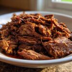 Close-up of tender, shredded Instant Pot Beef Barbacoa glistening with sauce in a white bowl.