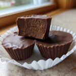 Three rich, dark chocolate Keto Chocolate Peanut Butter Fat Bombs in white paper liners, one cut open to show the dense filling.