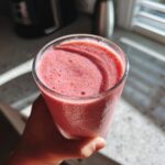 A hand holding a glass filled with a thick, pink Keto Strawberry Cheesecake Smoothie, backlit by sunlight.