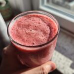 Close-up of a hand holding a glass filled with a frothy, pink Keto Strawberry Cheesecake Smoothie near a window.