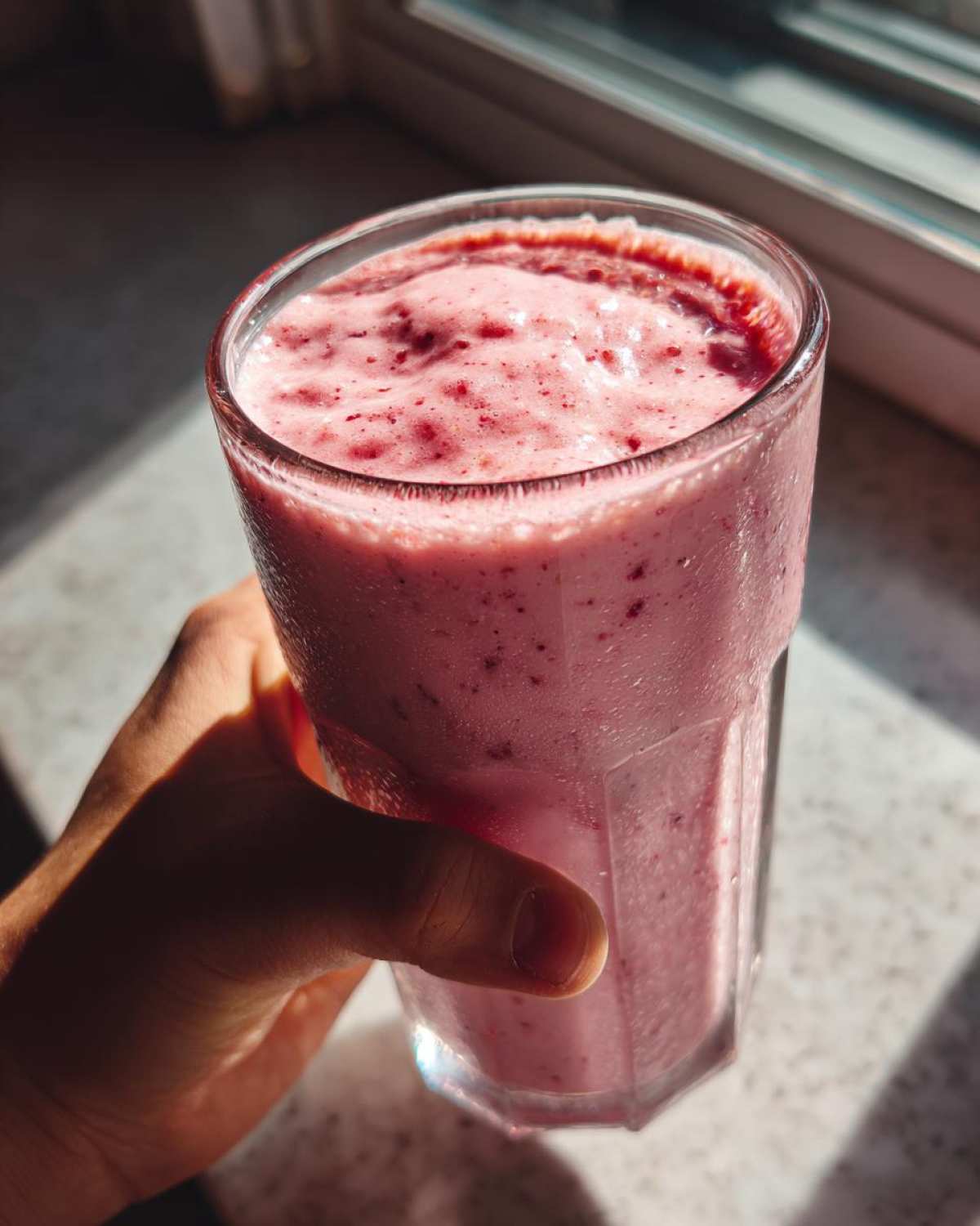 A hand holding a tall glass filled with a thick, pink Keto Strawberry Cheesecake Smoothie near a sunlit window.