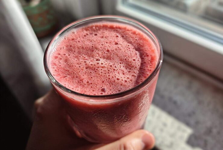 Close-up of a hand holding a glass filled with a frothy, pink Keto Strawberry Cheesecake Smoothie near a window.