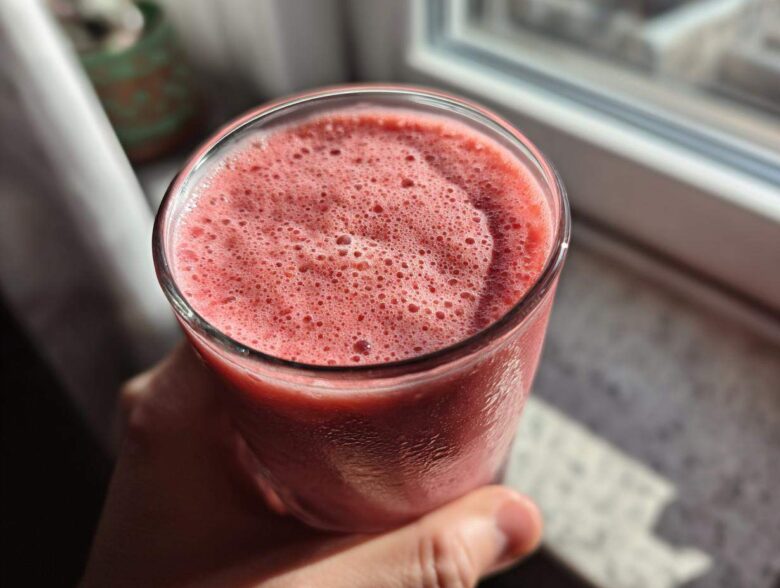 Close-up of a hand holding a glass filled with a frothy, pink Keto Strawberry Cheesecake Smoothie near a window.