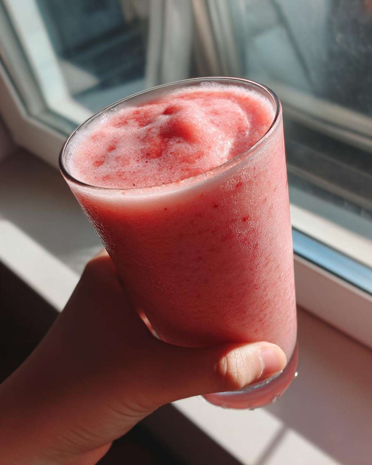 A hand holding a tall glass filled with a thick, pink Keto Strawberry Cheesecake Smoothie near a bright window.