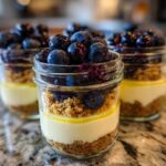 Three layered Lemon Blueberry Cheesecake Jars featuring graham cracker crust, creamy filling, and fresh blueberries on top.