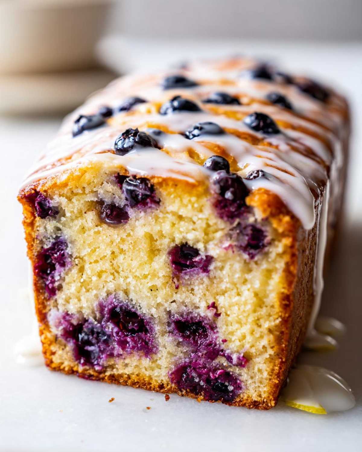 Close-up of a moist Lemon Blueberry Yogurt Loaf topped with a white lemon glaze and fresh blueberries.