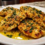 Four fillets of Lemon Garlic Butter Tilapia swimming in a pool of bright yellow garlic butter sauce, topped with fresh parsley.