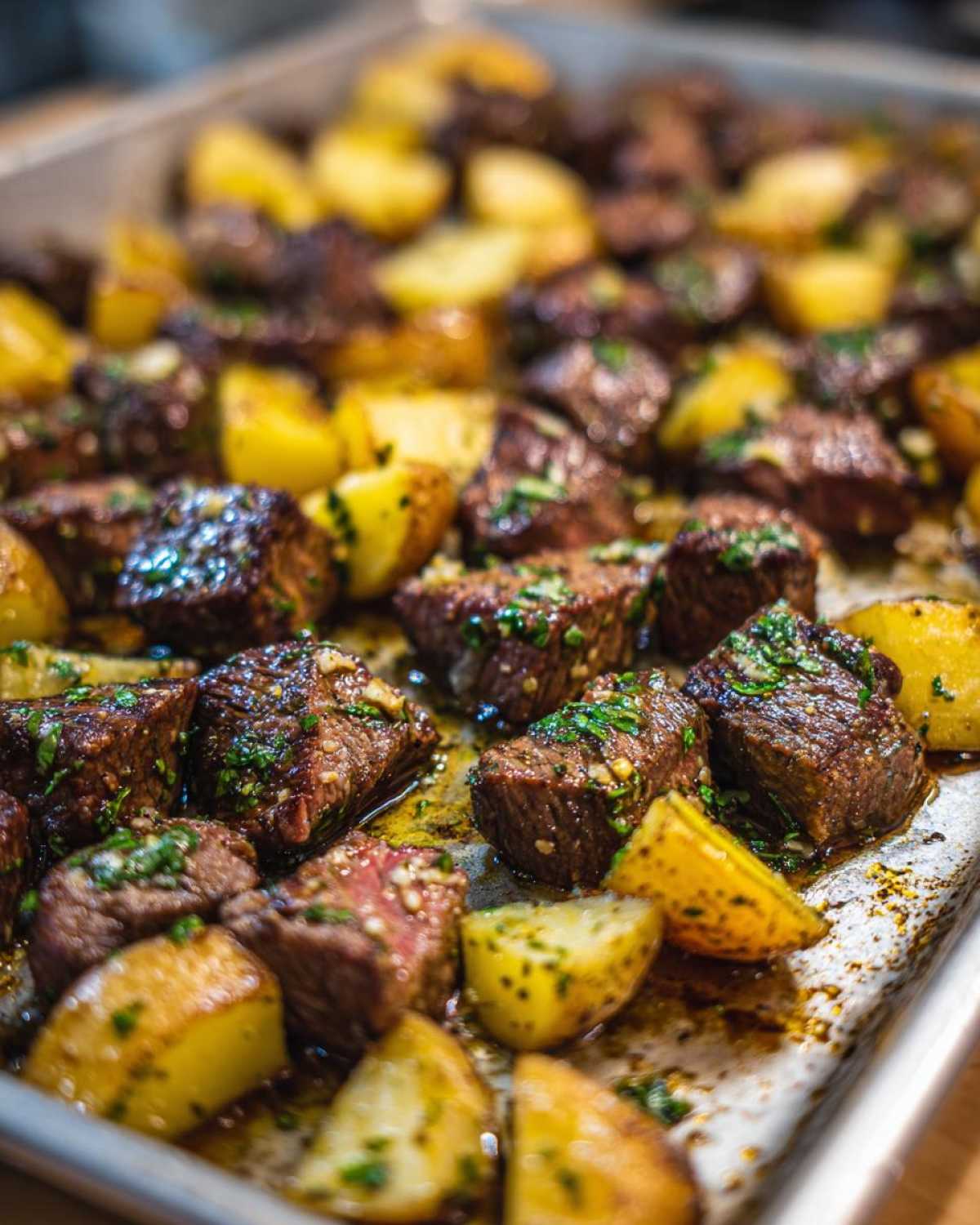 Close-up of juicy, browned steak bites mixed with roasted potatoes, coated in garlic butter sauce and parsley, fresh from the pan.