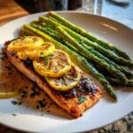 A perfectly cooked fillet of One-Pan Lemon Garlic Salmon topped with roasted lemon slices and parsley, served next to bright green asparagus.