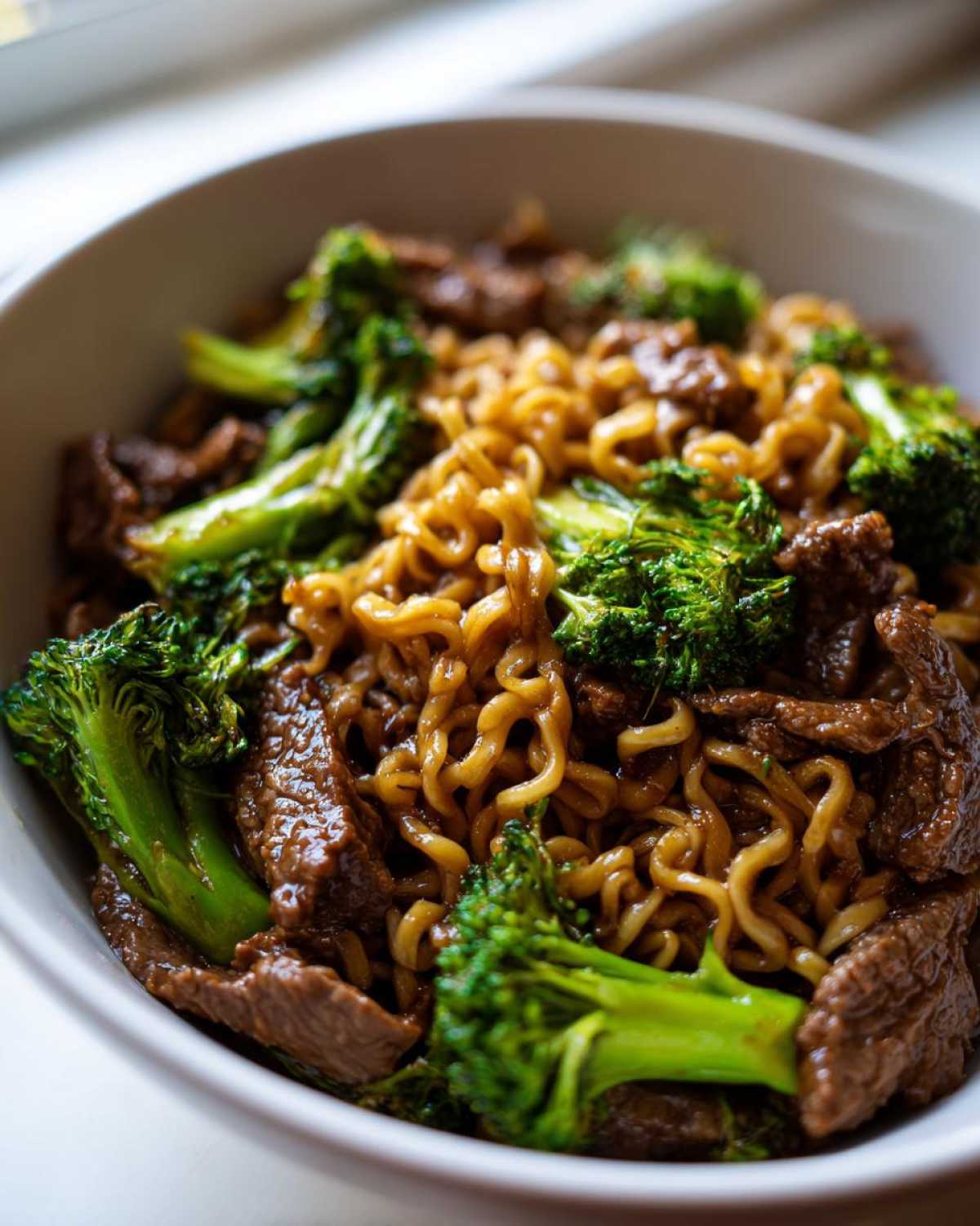 A close-up view of a white bowl filled with One-Pot Beef and Broccoli Ramen Noodles, featuring glossy noodles, tender beef strips, and bright green broccoli florets.