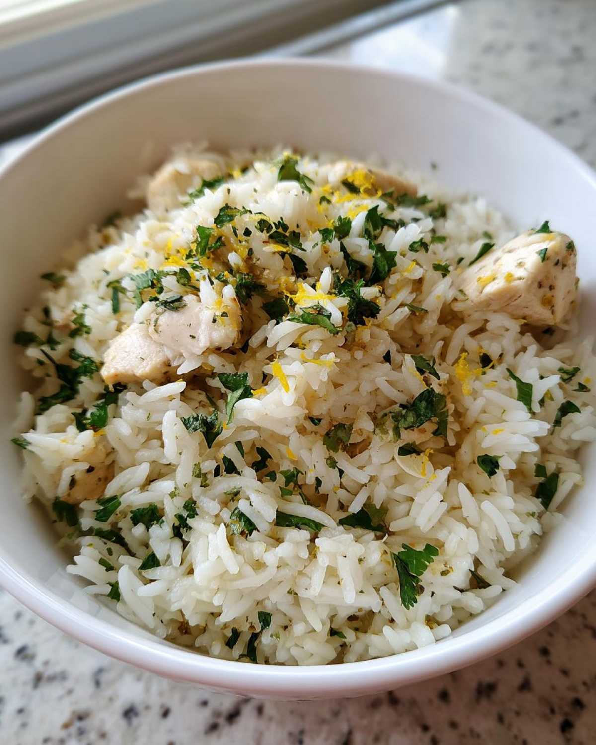 A white bowl filled with fluffy white rice, chunks of chicken, and topped generously with fresh parsley and lemon zest from the One-Pot Chicken and Rice with Lemon and Herbs.