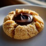 A single, freshly baked Peanut Butter Blossom Cookie with a swirl of melted chocolate center, sitting on a white plate.
